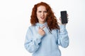 Confused ginger girl student pointing finger at empty phone screen, showing smartphone app on display, white background