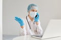 Confused helpless woman doctor wearing gown, surgical mask, medical cap and gloves, posing in front of notebook, at desk, talking