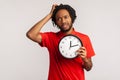 Confused man with dreadlocks wearing red T-shirt, rubbing his head holding in hands big wallclock, has no time , worried about Royalty Free Stock Photo