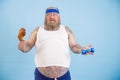 Confused man with overweight chooses between chicken leg and dumbbell on light blue background Royalty Free Stock Photo