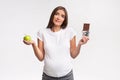 Confused Pregnant Lady Holding Apple And Chocolate Standing In Studio