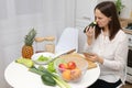 Confused puzzled Caucasian brown haired woman sitting at kitchen table with fruit and vegetables smelling not fresh cucumber from Royalty Free Stock Photo