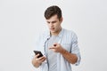 Confused puzzled male with dark hair wearing blue shirt chatting, using phone and pointing with forefinger at screen of