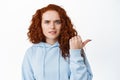 Confused redhead curly girl showing something strange, pointing right and frowning doubtful, standing hesitant and