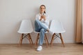 Confused sad attractive female with ponytail sitting on chair against light wall, having funny facial expression, woman wearing Royalty Free Stock Photo