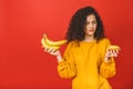 Confused sad upset curly woman can`t make a choice between healthy and unhealthy food, she is holding fruits and donut on her Royalty Free Stock Photo