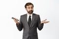 Confused salesman holding phone and shrugging, looking unaware and clueless, standing in grey suit over white background