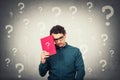 Confused student guy looks down upset, holding a book with question mark and different interrogation symbols around head. Concept Royalty Free Stock Photo