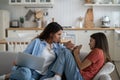 Caring attentive woman with laptop on knees sits on sofa of apartment near teen girl with smartphone