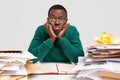 Confused tired black male student keeps hands under chin, wears spectacles and green jumper, looks in bewilderment Royalty Free Stock Photo