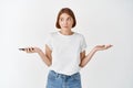 Confused woman holding smartphone and shrugging unaware, standing clueless against white background Royalty Free Stock Photo