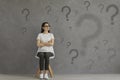 Confused woman sitting on chair and looking at lots of question marks on grey background