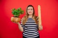 Confused young long-haired plump woman in striped shirt holding green flower pot and long french baguette in red studio Royalty Free Stock Photo