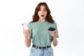 Confused young woman holding plastic credit card and smartphone, open mouth and looking with indecisive worried face at