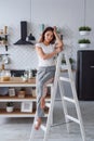 Positive beautiful young woman on a white wooden stepladder on the background of a stylish modern kitchen. Royalty Free Stock Photo