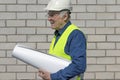 A construction man examines a sheet of paper. He is wearing a vest and helmet. Portrait of a male builder on a white brick backgro