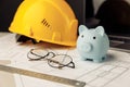 Construction safety helmet and piggy bank with blueprint, glasses and laptop on table. Building theme Royalty Free Stock Photo