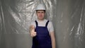 Builder in blue overalls puts on white hard hat and shows thumbs up. Royalty Free Stock Photo