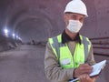 Construction worker dressed in medical mask, uniform and white hardhat stands with clipboard at metro tunnel Royalty Free Stock Photo