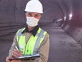 Construction worker dressed in medical mask, uniform and white hardhat stands with clipboard at metro tunnel Royalty Free Stock Photo