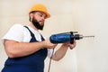 Construction worker, handyman and builder with blue electric drill drills a nail hole into wall of construction site.