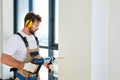 Construction worker making hole in white wall with electric drill, side view