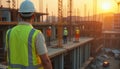 Construction worker observes site progress at sunset. Team of builders works hard, wearing safety vests, helmets. Construction Royalty Free Stock Photo