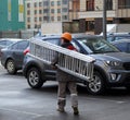 A construction worker in overalls and an orange hard hat carries a sliding ladder through the Parking lot