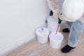 A construction worker in protective clothing and a hard hat mixes paint with a mixer in a bucket, against a white wall
