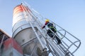 Construction worker in safety harness and hard hat climbing a fixed ladder on a cement silo at a small concrete batching plant. Royalty Free Stock Photo