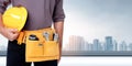 Construction worker with tool belt holding yellow hard hat standing in front of city skyline background, copy space Royalty Free Stock Photo