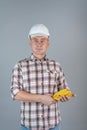 A construction worker in a white helmet, standing in casual clothes on a gray background and holding yellow protective gloves in Royalty Free Stock Photo