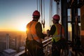 Construction workers adjusting equipment high above the city during a radiant sunset.