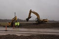Construction workers stand by while they watch two Caterpillar excavators dig utility line for new freeway overpass on ramp.