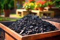 A container filled with biochar stands in a garden with green plants and colorful flowers in the background. Biochar increases