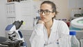 A contemplative young hispanic woman scientist with glasses in a lab coat in a laboratory setting with a microscope Royalty Free Stock Photo