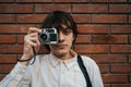 Contemplative young man peers through viewfinder of retro style camera, framed against red brick wall backdrop Royalty Free Stock Photo