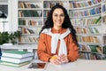 Contemporary online tutor sitting at table in library surrounded by shelves filled with books. Pretty woman wearing an Royalty Free Stock Photo