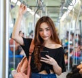 Content Asian female riding train and chatting on social media via smartphone while holding handrail and looking at camera.