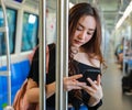 Content Asian female riding train and chatting on social media via smartphone while holding handrail and looking at phone screen.