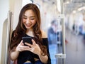 Content Asian female riding train and chatting on social media via smartphone while holding handrail and looking at phone screen.