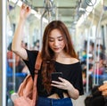 Content Asian female riding train and chatting on social media via smartphone while holding handrail and looking at screen.