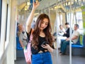 Content Asian female riding train and chatting on social media via smartphone while holding handrail and looking at screen of