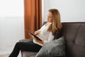 Content pregnant woman holding her tablet sitting on a couch in the living room at home