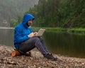 A contented bearded guy in a blue jacket with a hood sits on the bank of a river in the rain with a laptop in his lap. Freelancer