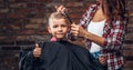 Contented cute preschooler boy shows thumbs up while getting a haircut. Children hairdresser with scissors and comb is