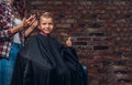 Contented cute preschooler boy shows thumbs up while getting a haircut. Children hairdresser with scissors and comb is