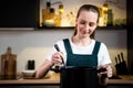 Contented, happy young housewife woman in an apron holds a spoon of a pot of soup, tastes soup from a pot. The concept