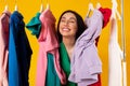 Contented shopaholic woman peeking out between clothes hanging in rail and smiling with closed eyes, yellow background