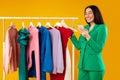 Contented young shopaholic woman with cellphone shopping while standing near clothing rail, choosing trendy clothes